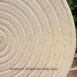 May include: A circular ceramic plate with a light beige base colour and dark brown speckles throughout. The surface features concentric circular grooves radiating from the centre. The text "Amount of specks may vary" is printed at the bottom.