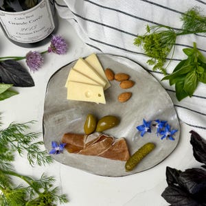 Puede incluir: Un plato de cerámica gris con rodajas de queso, almendras, aceitunas, prosciutto y un pepinillo. Flores azules y hierbas frescas rodean el plato. Una botella de sidra es visible en el fondo.