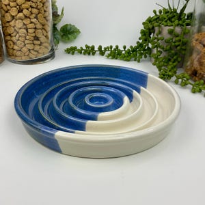 May include: A round ceramic slow feeder bowl featuring a series of concentric raised rings in the centre. The bowl is finished with a two-tone glaze, displaying a deep blue colour on one side and a smooth, creamy off-white on the other side.