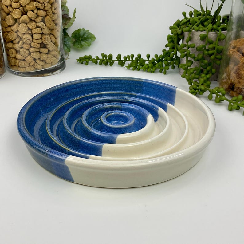 May include: A round ceramic slow feeder bowl featuring a series of concentric raised rings in the centre. The bowl is finished with a two-tone glaze, displaying a deep blue colour on one side and a smooth, creamy off-white on the other side.