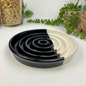 May include: A circular ceramic slow feeder bowl featuring a concentric ring design. The bowl is divided into two distinct halves, with one side finished in a glossy black glaze and the other side finished in a smooth off-white cream glaze.