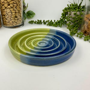 May include: A round ceramic slow feeder bowl featuring a series of concentric raised rings. The bowl is finished in a two-tone glaze, with one half in a vibrant lime green and the other half in a deep navy blue, creating a distinct colour split.