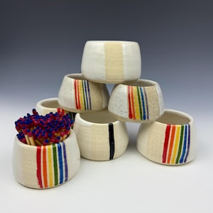 May include: A collection of handmade ceramic match holders. The holders are white with a textured beige stripe and rainbow stripes. One holder contains matches with blue tips. The holders are stacked in a pyramid shape.