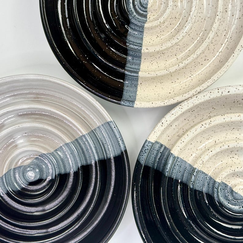 May include: Three ceramic plates featuring a circular ridged texture. Each plate is divided into two distinct sections, one in solid black and the other in a speckled off-white, separated by a band of grey glaze that follows the concentric rings.