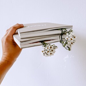 K&ouml;nnte beinhalten: Ein Stapel von drei grauen Hardcover-Journalen mit wei&szlig;em Text auf dem vorderen Umschlag. Die Journale werden in einer Hand gehalten, in die kleine wei&szlig;e Blumen gesteckt sind.