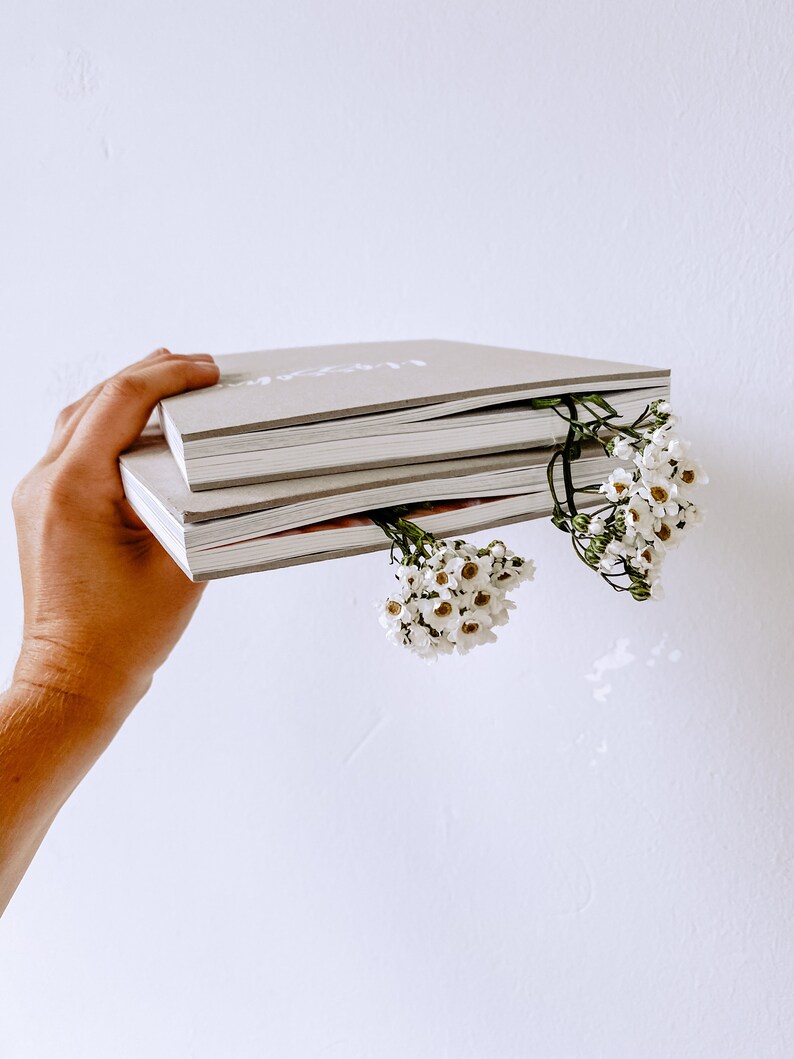K&ouml;nnte beinhalten: Ein Stapel von drei grauen Hardcover-Journalen mit wei&szlig;em Text auf dem vorderen Umschlag. Die Journale werden in einer Hand gehalten, in die kleine wei&szlig;e Blumen gesteckt sind.