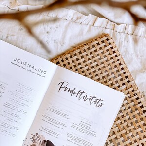 K&ouml;nnte beinhalten: Nahaufnahme eines ge&ouml;ffneten Buches mit den Titeln "Journaling" und "Produktivit&auml;ts" auf den Seiten. Das Buch ist auf einer Seite mit einer Aquarell-Illustration eines Baumes und einer Seite mit Text &uuml;ber Journaling und Produktivit&auml;tstipps ge&ouml;ffnet.