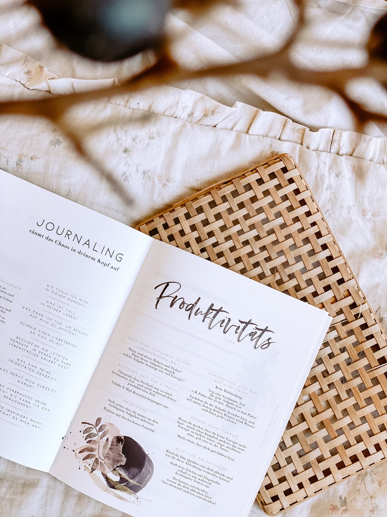 K&ouml;nnte beinhalten: Nahaufnahme eines ge&ouml;ffneten Buches mit den Titeln "Journaling" und "Produktivit&auml;ts" auf den Seiten. Das Buch ist auf einer Seite mit einer Aquarell-Illustration eines Baumes und einer Seite mit Text &uuml;ber Journaling und Produktivit&auml;tstipps ge&ouml;ffnet.