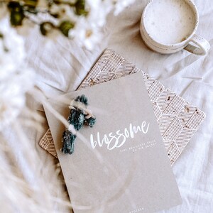 K&ouml;nnte beinhalten: Ein braunes Papierjournal mit dem Titel "blossom" in wei&szlig;er Schrift. Der Text darunter lautet "eine bewusste reise zu dir selbst" und "Simone Stocker". Das Journal liegt auf einer wei&szlig;en Oberfl&auml;che mit einer wei&szlig;en Tasse Kaffee und einem Strau&szlig; wei&szlig;er Blumen.