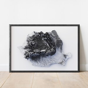 May include: Framed grayscale relief map of Romania, showcasing the country's topography. The artwork is encased in a black frame, set against a white background, and rests on a wooden floor.