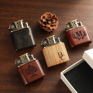 May include: Five wooden lighters with brass detailing are arranged on a wooden surface. Each lighter features a unique engraved design, including a dollar sign, the letter "M" with antlers, the letter "B", and a spade with the word "CHASSE". A pinecone is in the center.
