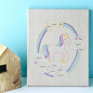 May include: A pink unicorn embroidered on a beige canvas with a rainbow and clouds. The unicorn has a gold crown and a rainbow mane and tail. The clouds have happy faces.
