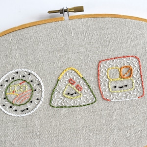 May include: Embroidered sushi design on a beige linen fabric stretched in a wooden embroidery hoop. The design features three different types of sushi: a maki roll, a nigiri, and a sashimi.