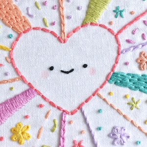 May include: Hand-stitched embroidery art featuring a smiling heart with rosy cheeks, surrounded by colorful rays and floral accents. The design incorporates various embroidery stitches in shades of pink, yellow, green, and orange on a white fabric background. A whimsical and cheerful design.