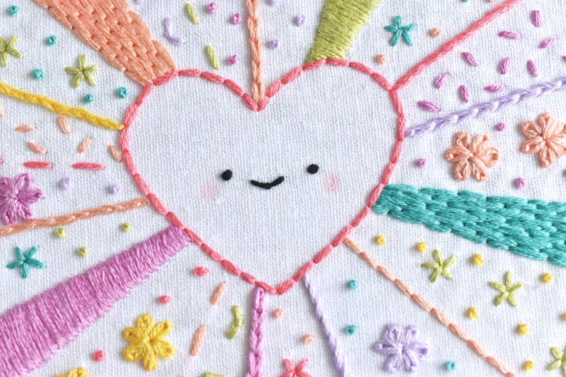 May include: Hand-stitched embroidery art featuring a smiling heart with rosy cheeks, surrounded by colorful rays and floral accents. The design incorporates various embroidery stitches in shades of pink, yellow, green, and orange on a white fabric background. A whimsical and cheerful design.
