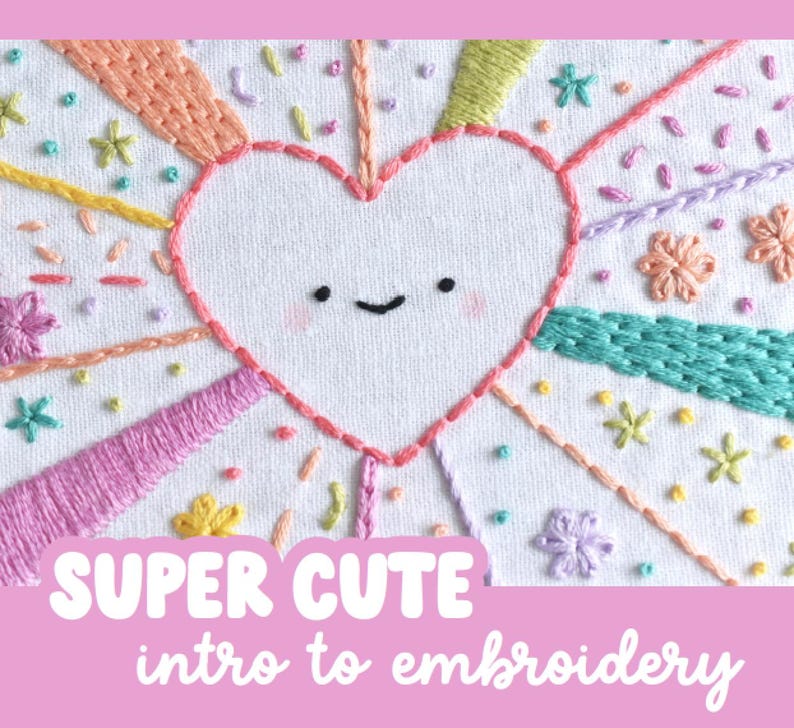 May include: Embroidered heart design with a smiling face, surrounded by colorful rays and floral accents. The text "SUPER CUTE intro to embroidery" is displayed in a pink banner at the bottom. The embroidery is on a white fabric background.