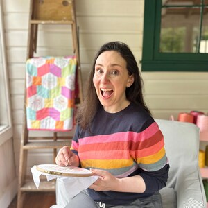 May include: A person is shown smiling while doing embroidery. They are wearing a navy blue sweater with horizontal stripes of pink, orange, yellow, and red. A wooden embroidery hoop is in their hands. A colorful quilt and a wooden ladder are in the background.
