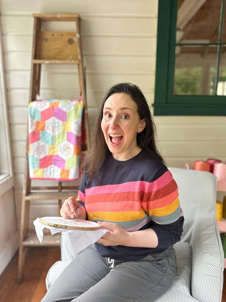 May include: A person is shown smiling while doing embroidery. They are wearing a navy blue sweater with horizontal stripes of pink, orange, yellow, and red. A wooden embroidery hoop is in their hands. A colorful quilt and a wooden ladder are in the background.