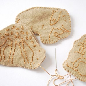 May include: Three beige felt shapes with gold embroidery. The shapes are stitched together with a gold thread. The shapes are in the form of leaves with different designs.