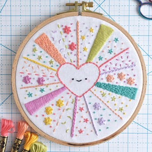 May include: Embroidered heart design in a wooden hoop. The heart is surrounded by colorful rays and floral accents. Embroidery floss in various colors is visible, along with a pair of unicorn-shaped scissors.