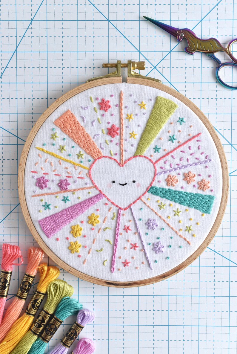 May include: Embroidered heart design in a wooden hoop. The heart is surrounded by colorful rays and floral accents. Embroidery floss in various colors is visible, along with a pair of unicorn-shaped scissors.