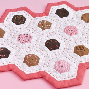 Peut inclure: Un dessous de plat hexagonal matelassé rose et blanc avec des motifs brodés en brun, rose et blanc de différents biscuits avec des visages.