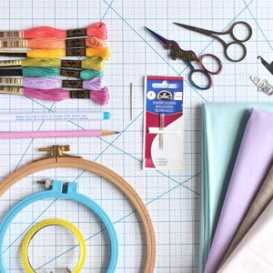 May include: An overhead shot of embroidery supplies, including colorful thread skeins, embroidery hoops, fabric, scissors, needles, and a pencil. The thread colors range from pink to yellow. The fabric is in shades of blue, purple, and white.