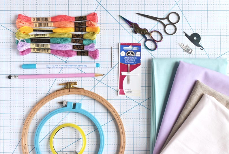May include: An overhead shot of embroidery supplies, including colorful thread skeins, embroidery hoops, fabric, scissors, needles, and a pencil. The thread colors range from pink to yellow. The fabric is in shades of blue, purple, and white.
