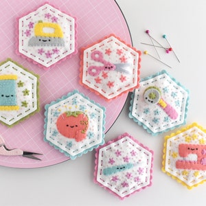 Op de afbeelding: Zeven viltnaaldenkussens in de vorm van zeshoeken, elk met een ander naai-thema. De naaldenkussens zijn versierd met kleurrijke borduurwerk en hebben schattige gezichten. De naaldenkussens zijn gerangschikt op een roze snijmat.