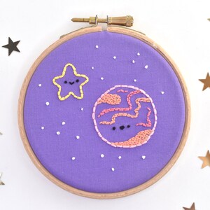 May include: Purple embroidery hoop with a pink and orange planet with a white star and white dots on a purple background.