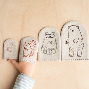 以下が含まれることがあります： ベージュの生地でできた指人形が4つ。刺繍で動物のデザインが施されています。人形は小さい順に並んでいます。動物は、ネズミ、リス、アライグマ、クマです。