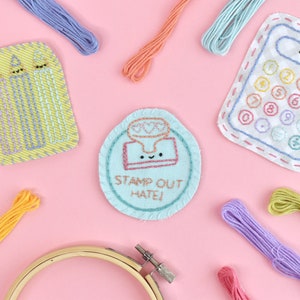 May include: A set of three embroidered patches featuring a calculator, a box of pencils, and a stamp with the text "Stamp Out Hate!"  The patches are surrounded by colorful embroidery floss.