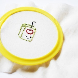 May include: Two embroidery hoops with hand-stitched designs. The first hoop features a green juice box with a red apple and a straw. The second hoop features a yellow lemon with a blue stem and a happy face.
