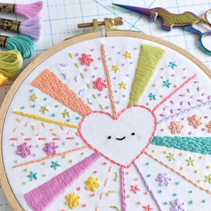 May include: A white embroidery hoop with a colorful embroidered design of a heart with a smiling face surrounded by radiating lines and flowers. The hoop is on a white gridded surface with colorful embroidery floss spools.