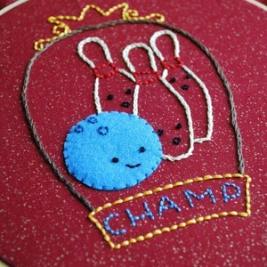 May include: Embroidered hoop art featuring a blue bowling ball with a smiling face and three white bowling pins with red stripes. The hoop is decorated with a gold crown and the word "CHAMP" in gold thread.