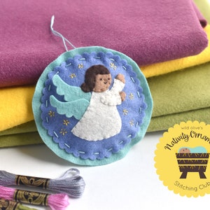 May include: A handmade felt angel Christmas ornament with a blue background, white dress, and teal wings. The ornament is decorated with gold stitching and stars. The background includes purple, yellow, and green felt fabric. The image also includes embroidery floss and a logo that says "Nativity Ornament Stitching Club."
