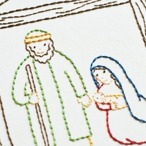 May include: Embroidered image of the nativity scene with Joseph, Mary, and baby Jesus. Joseph is wearing a green robe and a yellow hat. Mary is wearing a blue robe and a red dress. Baby Jesus is wrapped in a yellow blanket.