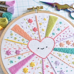 May include: Embroidered hoop art featuring a white heart with a smiling face, surrounded by colorful rays and floral accents. Embroidery floss in various pastel colors and a pair of rainbow scissors are also visible. A craft project.