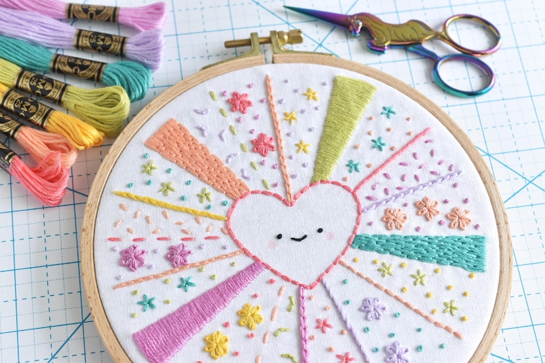 May include: Embroidered hoop art featuring a white heart with a smiling face, surrounded by colorful rays and floral accents. Embroidery floss in various pastel colors and a pair of rainbow scissors are also visible. A craft project.