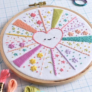 May include: Hand-stitched embroidery art in a wooden hoop. The design features a smiling heart at the center, with colorful rays and floral accents. Embroidery floss in various colors and rainbow scissors are also visible.