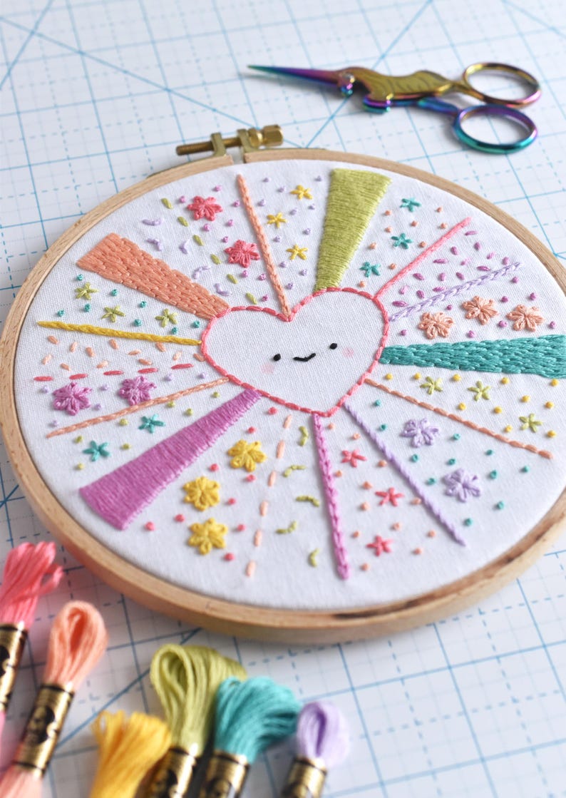 May include: Hand-stitched embroidery art in a wooden hoop. The design features a smiling heart at the center, with colorful rays and floral accents. Embroidery floss in various colors and rainbow scissors are also visible.