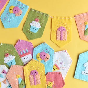 May include: A colorful felt birthday banner with various appliqued designs, including cupcakes, presents, balloons, and a birthday cake. The banner is strung together with a white string and features a light blue tag with the number one and a candle.