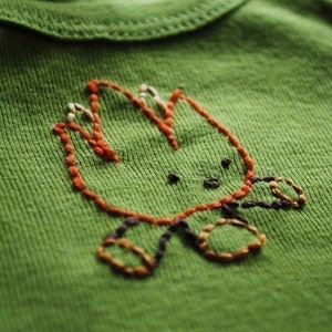 May include: A green t-shirt with an embroidered campfire design in orange, brown, and white thread.