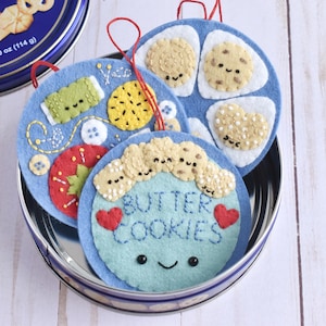 May include: A selection of felt ornaments in a tin. The ornaments are blue, decorated with embroidered cookie designs, and the text "BUTTER COOKIES". Each ornament has a red loop for hanging.