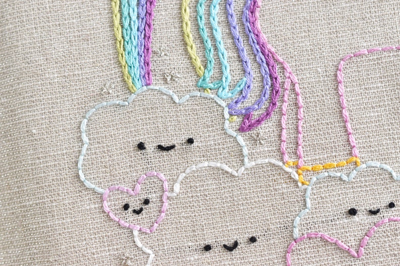 May include: Embroidered design of a rainbow with clouds and hearts. The clouds and hearts have smiling faces. The rainbow is made of multiple strands of yarn in different colors.