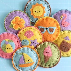 May include: Nine felt circles with different summer-themed designs, including a sun, sunglasses, a flamingo, a seashell, a starfish, a beach ball, a sailboat, a coconut, and an ice cream cone. Each circle has a stitched border and a cute face.