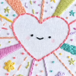 May include: Embroidered heart with a smiling face, surrounded by colorful rays and floral accents. The design features pink, orange, yellow, green, and purple thread on a white fabric background. A cheerful, handmade craft.