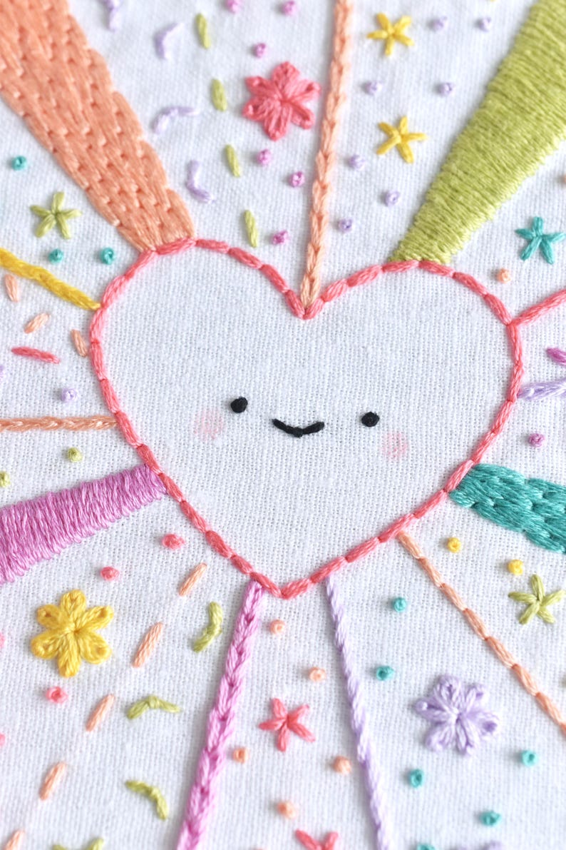 May include: Embroidered heart with a smiling face, surrounded by colorful rays and floral accents. The design features pink, orange, yellow, green, and purple thread on a white fabric background. A cheerful, handmade craft.