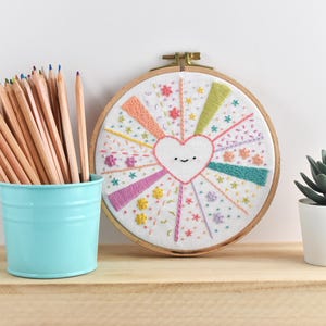 May include: A handmade embroidery hoop art piece with a smiling heart design, surrounded by colorful rays and floral accents. A light blue bucket holds a collection of pencils. A small potted succulent sits nearby.
