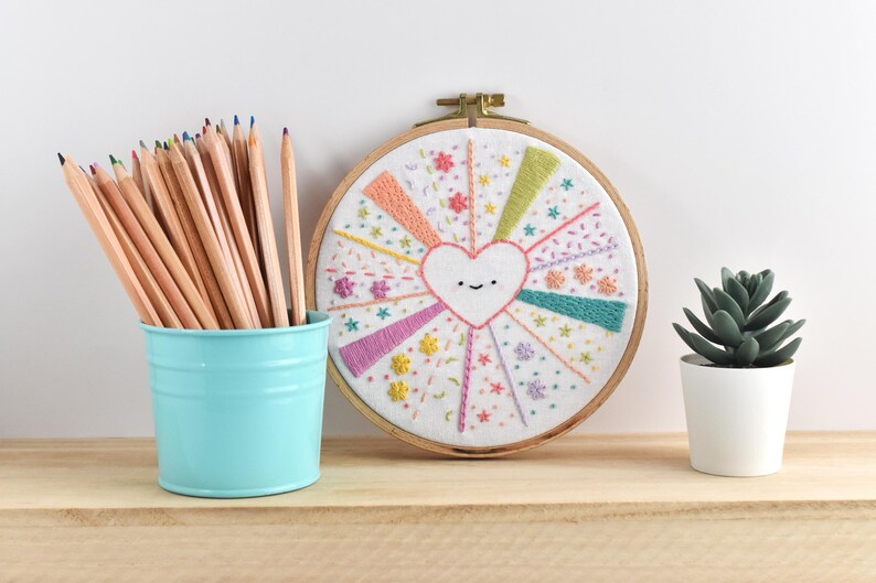 May include: A handmade embroidery hoop art piece with a smiling heart design, surrounded by colorful rays and floral accents. A light blue bucket holds a collection of pencils. A small potted succulent sits nearby.